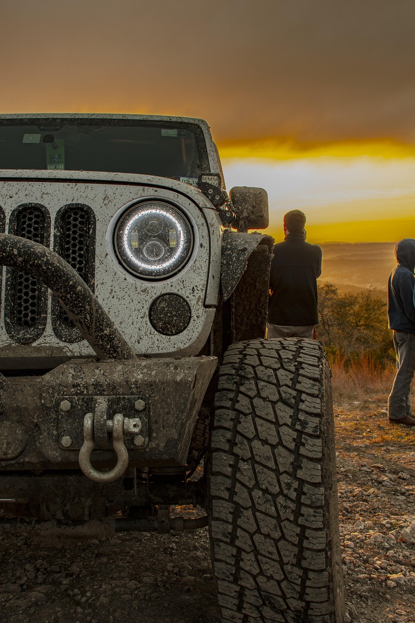 off roadiing in moab sunset Close-up of a muddy Jeep Wrangler parked on a rocky overlook near Moab, Utah, with golden desert light and two people watching the sunset.
