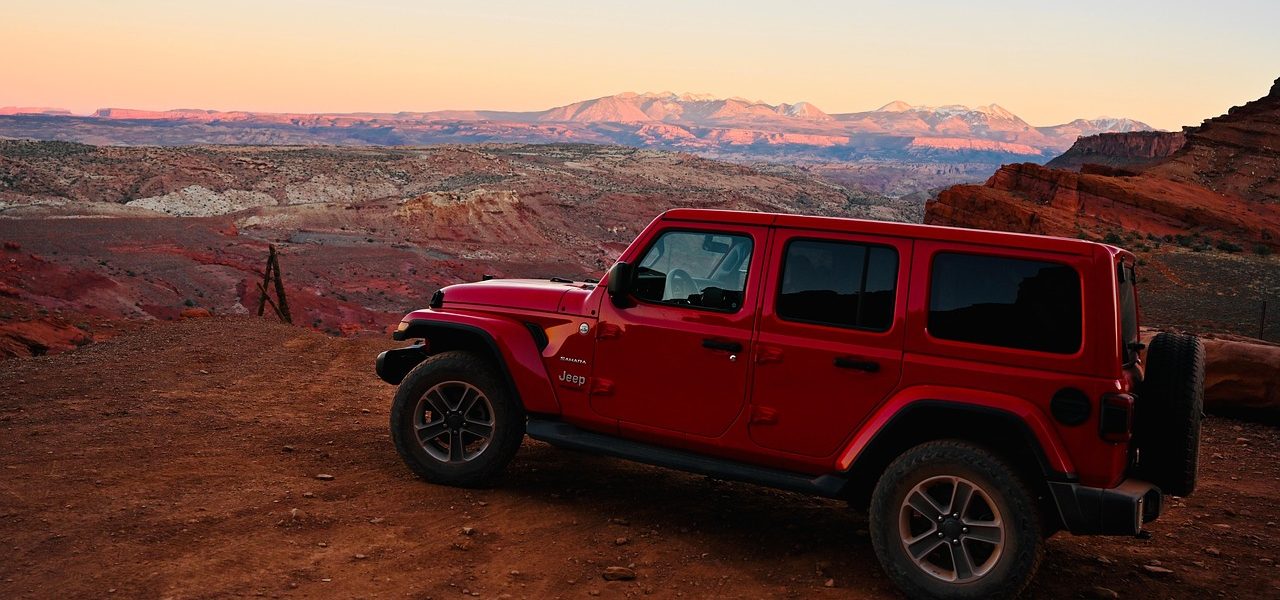 Red Jeep Wrangler parked on a canyon overlook near Moab, Utah, with distant La Sal Mountains glowing under sunset light.