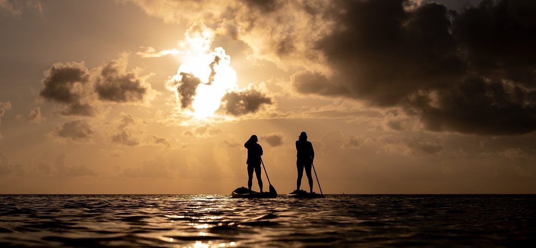 Two paddleboarders silhouetted against a glowing desert sunset, standing calmly on still water beneath dramatic clouds.