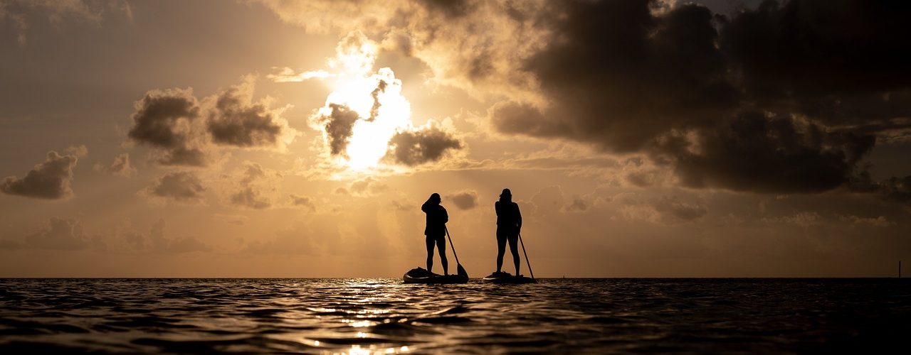 Two paddleboarders silhouetted against a glowing desert sunset, standing calmly on still water beneath dramatic clouds