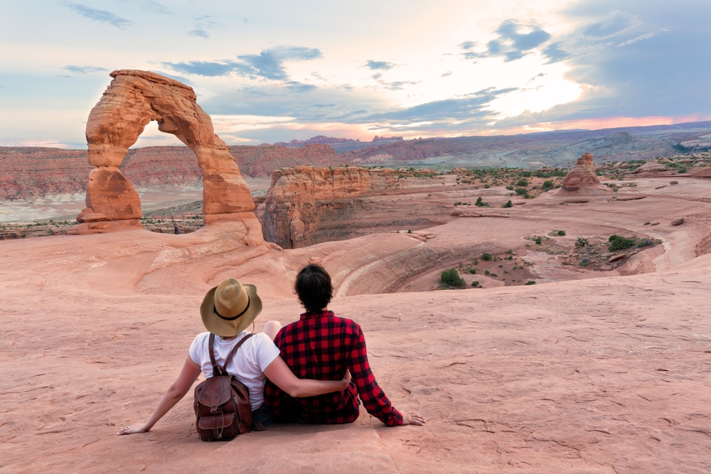 A couple sits arm in arm on a sandstone ledge, gazing across a canyon at Delicate Arch silhouetted against the sunset sky.