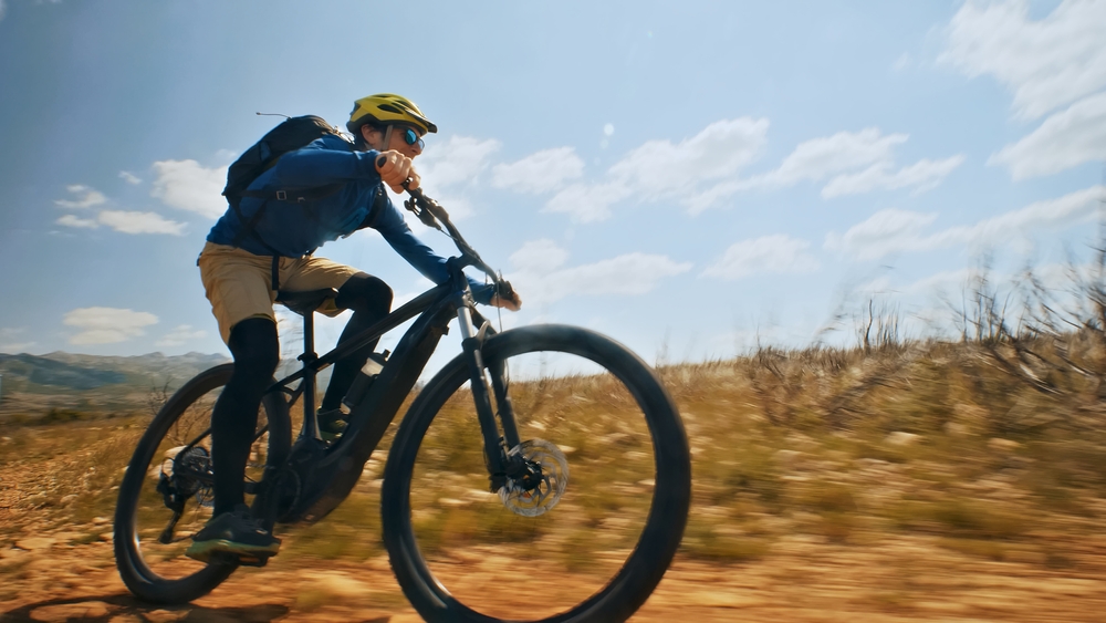 A cyclist in a yellow helmet rides a mountain e-bike along a dirt trail, motion blur emphasizing speed.