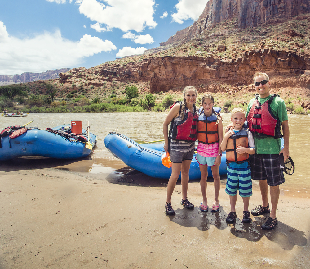 A family of four in life jackets poses on a sandy riverbank beside two rafts, with red canyon cliffs behind them.