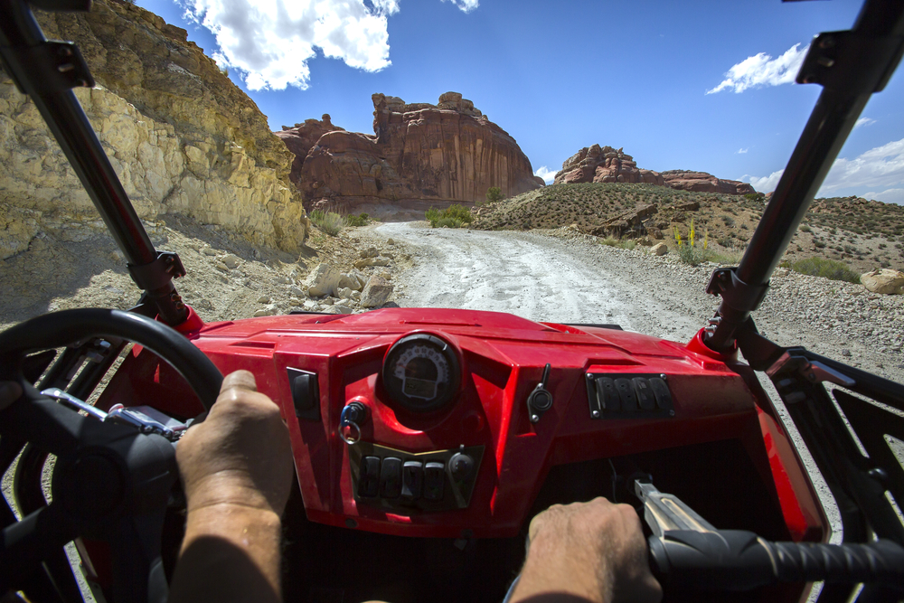 Driver's-eye view from inside a red UTV on a gravel trail, heading toward a red sandstone rock formation under a blue sky.