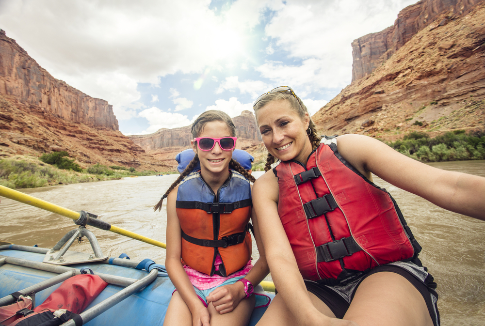 A mother and daughter in life jackets smile for a selfie while rafting down a river between red canyon walls.