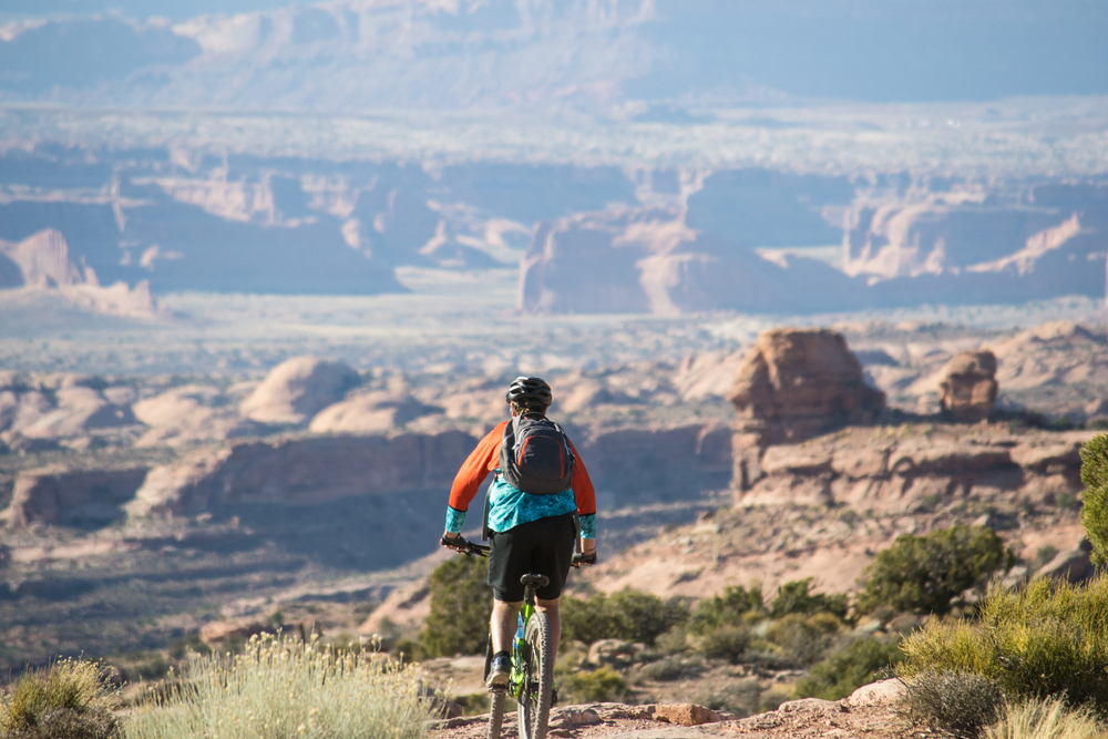 A mountain biker in an orange and teal jersey pauses on a trail overlooking a vast desert canyon dotted with rock towers.