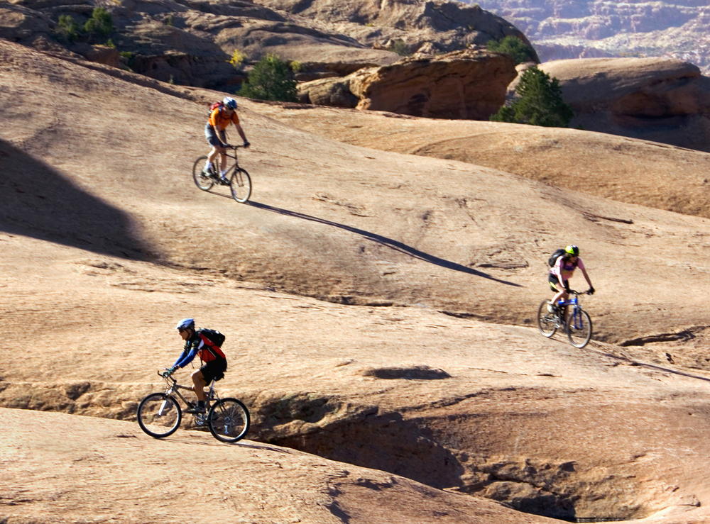 Three mountain bikers ride across red slickrock terrain with a canyon landscape in the background near Moab, Utah.