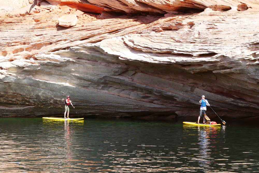 Two people stand on yellow paddleboards gliding through calm water beneath a striped sandstone rock overhang.