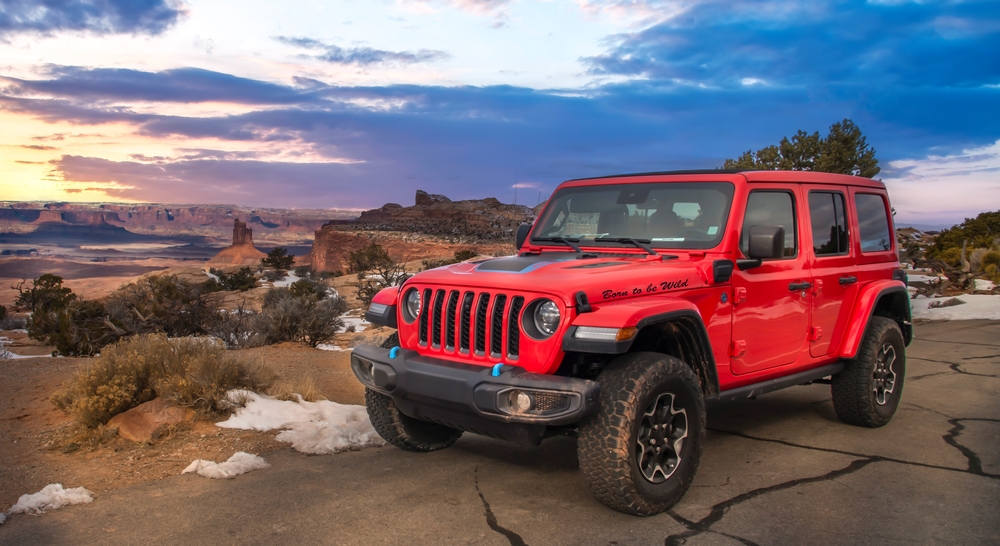 A red Jeep Wrangler is parked at a scenic overlook with patches of snow, overlooking a vast red rock canyon at dusk.