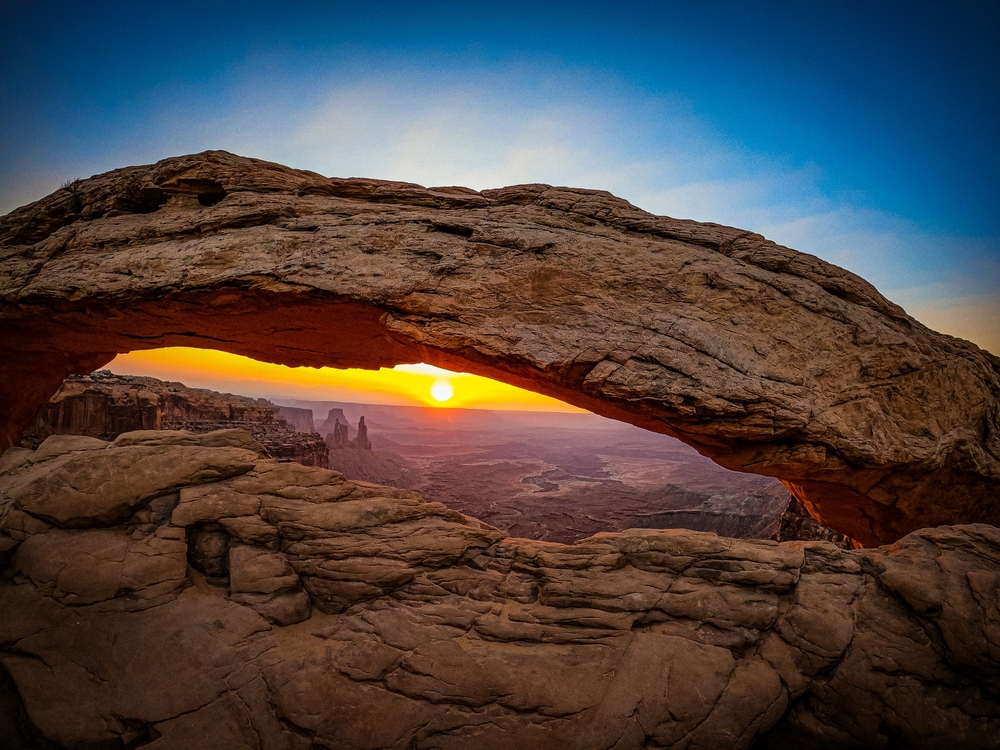 The sun rises over layered desert canyons as seen through the natural opening of Mesa Arch in Canyonlands National Park.