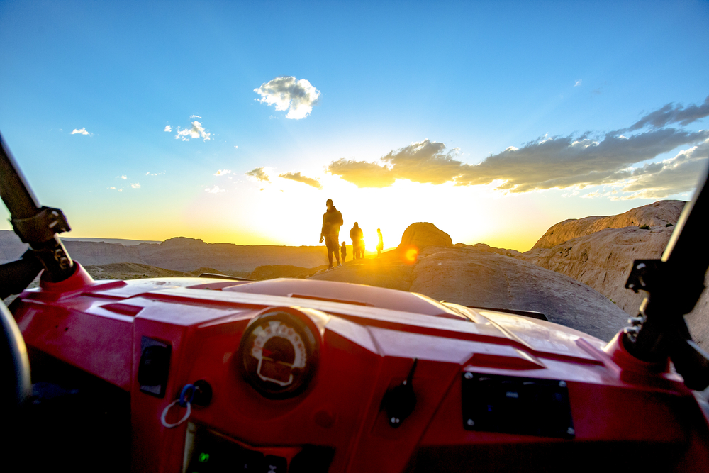 Silhouetted hikers stand on a rock ledge at sunset, viewed over the dashboard of an off-road UTV.