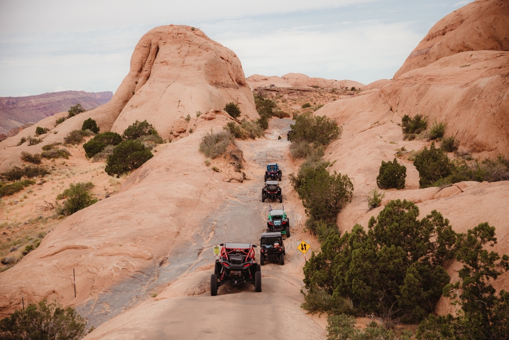 A line of off-road UTVs navigates a paved switchback trail between red rock formations in Moab, Utah.