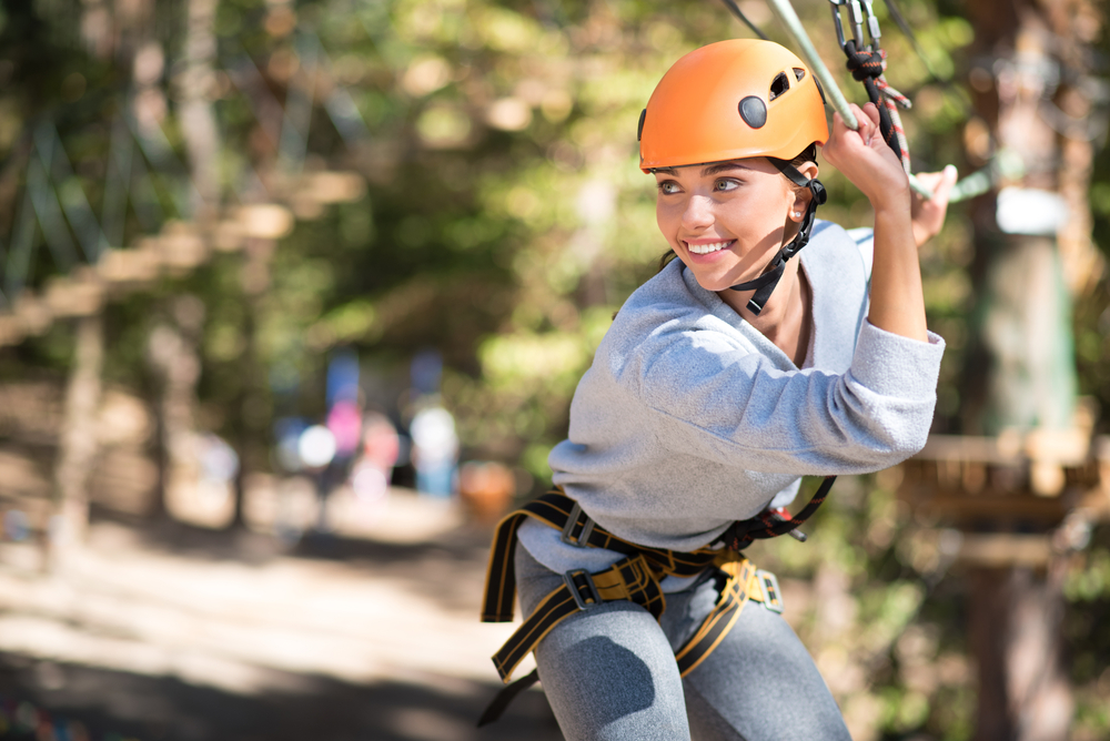 A smiling woman in an orange helmet and climbing harness prepares to zipline through a wooded ropes course.