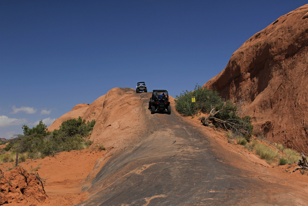 Two off-road UTVs climb a steep slickrock hill flanked by red rock formations and desert scrub near Moab, Utah.