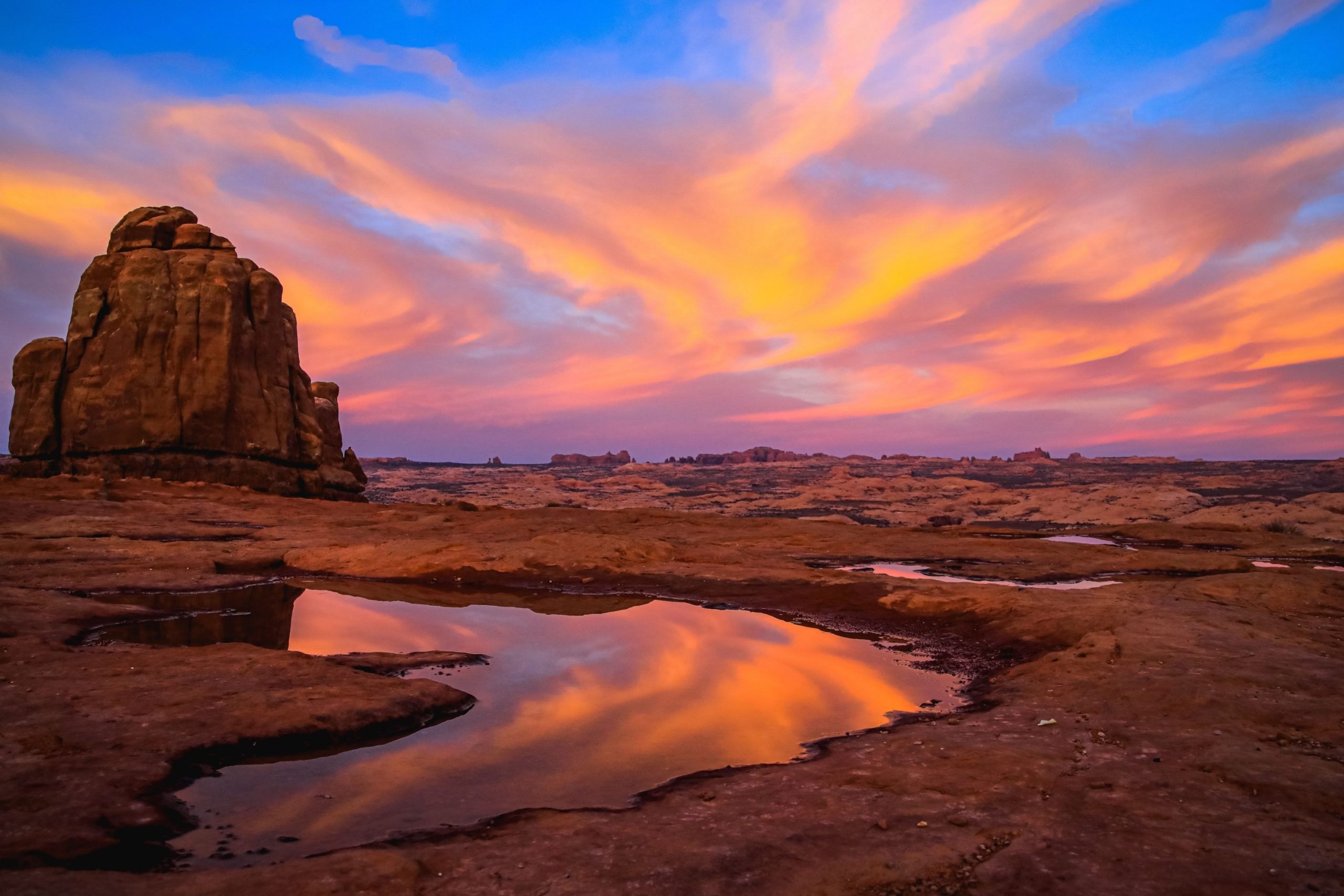 A vivid pink and orange sunset streaks across the sky above a sandstone rock formation in Arches National Park, reflected in a shallow pool on the desert floor.