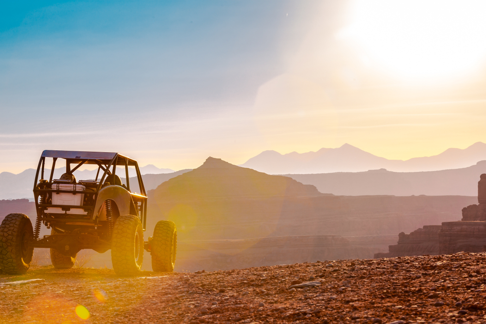 A dune buggy-style off-road vehicle sits atop a rocky overlook at sunset, with layered mountains and canyon silhouettes glowing in the hazy light.