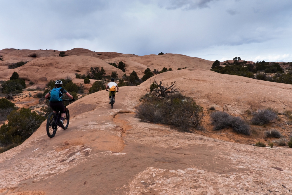Two mountain bikers ride across undulating red slickrock domes dotted with junipers under a cloudy sky near Moab, Utah