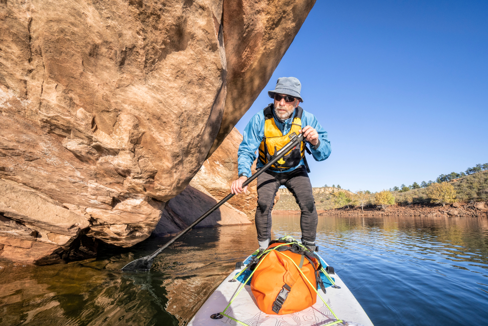 A paddleboarder in a life vest and bucket hat navigates his board past a towering sandstone overhang on a red rock lake.