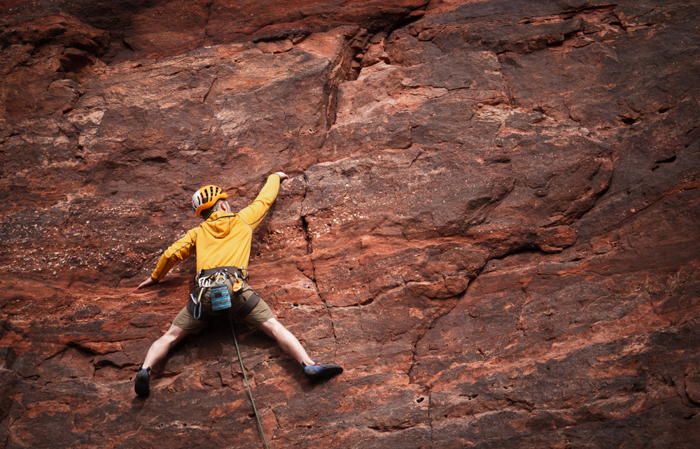 A rock climber in a yellow jacket and helmet stretches wide, reaching for a handhold on a steep red sandstone cliff face