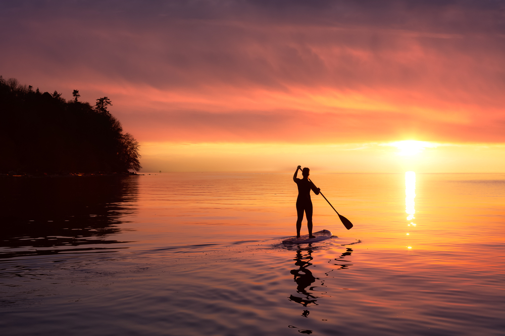 A paddleboarder is silhouetted against a vivid orange and pink sunset over calm water, paddle raised mid-stroke.