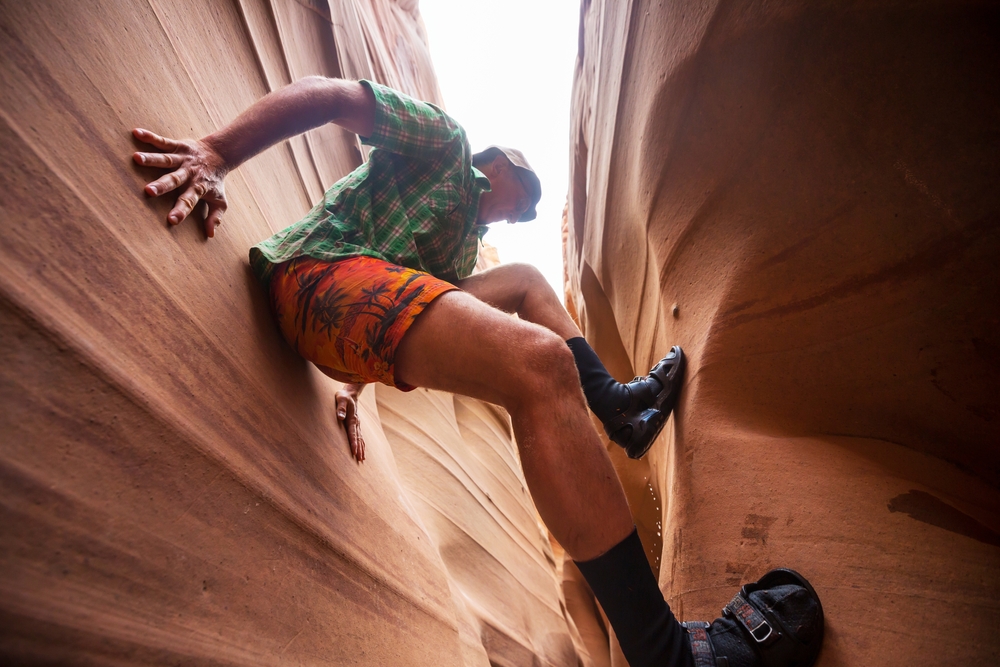 A hiker in a plaid shirt and patterned shorts climbs and braces between the narrow walls of a sandstone slot canyon.
