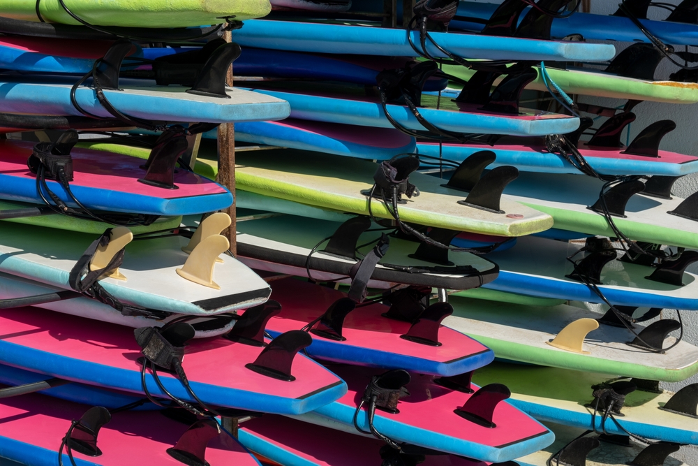 Rows of stacked stand-up paddleboards in blue, pink, and green line a rental rack, showing their fins and leash attachments.