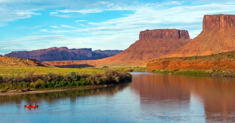 A small red raft drifts on the calm, reflective Colorado River beneath towering sandstone buttes near Moab, Utah