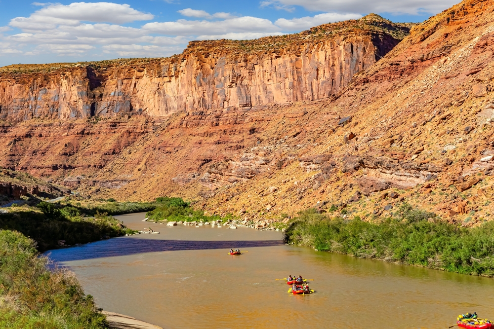 Rafts float down the muddy Colorado River, dwarfed by towering red and white sandstone cliffs near Moab