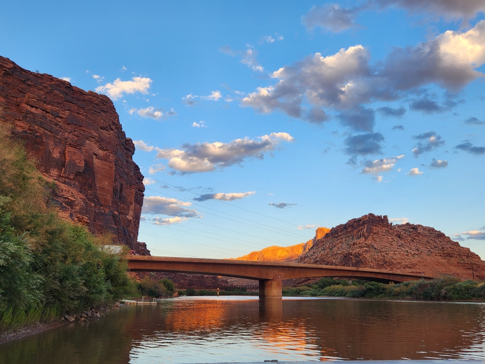 A road bridge spans the Colorado River near Moab, Utah, with red sandstone cliffs glowing at sunset. A popular put-in for paddleboarding trips.