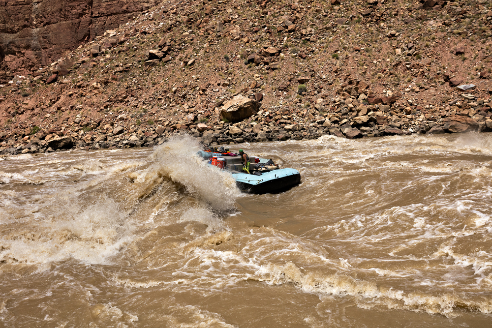 A raft plows through a large whitewater wave on a muddy river, with a rocky canyon slope in the background