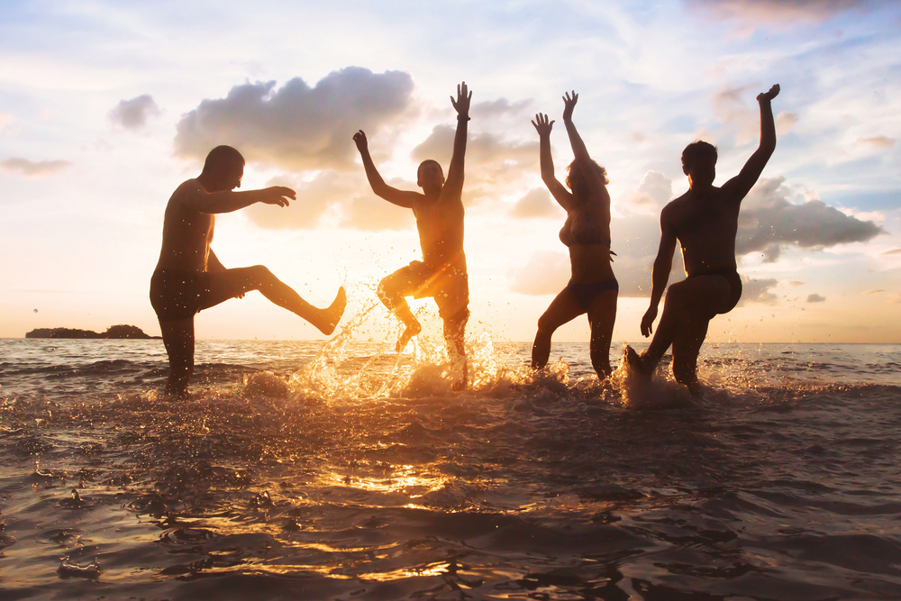 Four friends are silhouetted against a golden sunset, kicking up splashes of water as they play in the river.