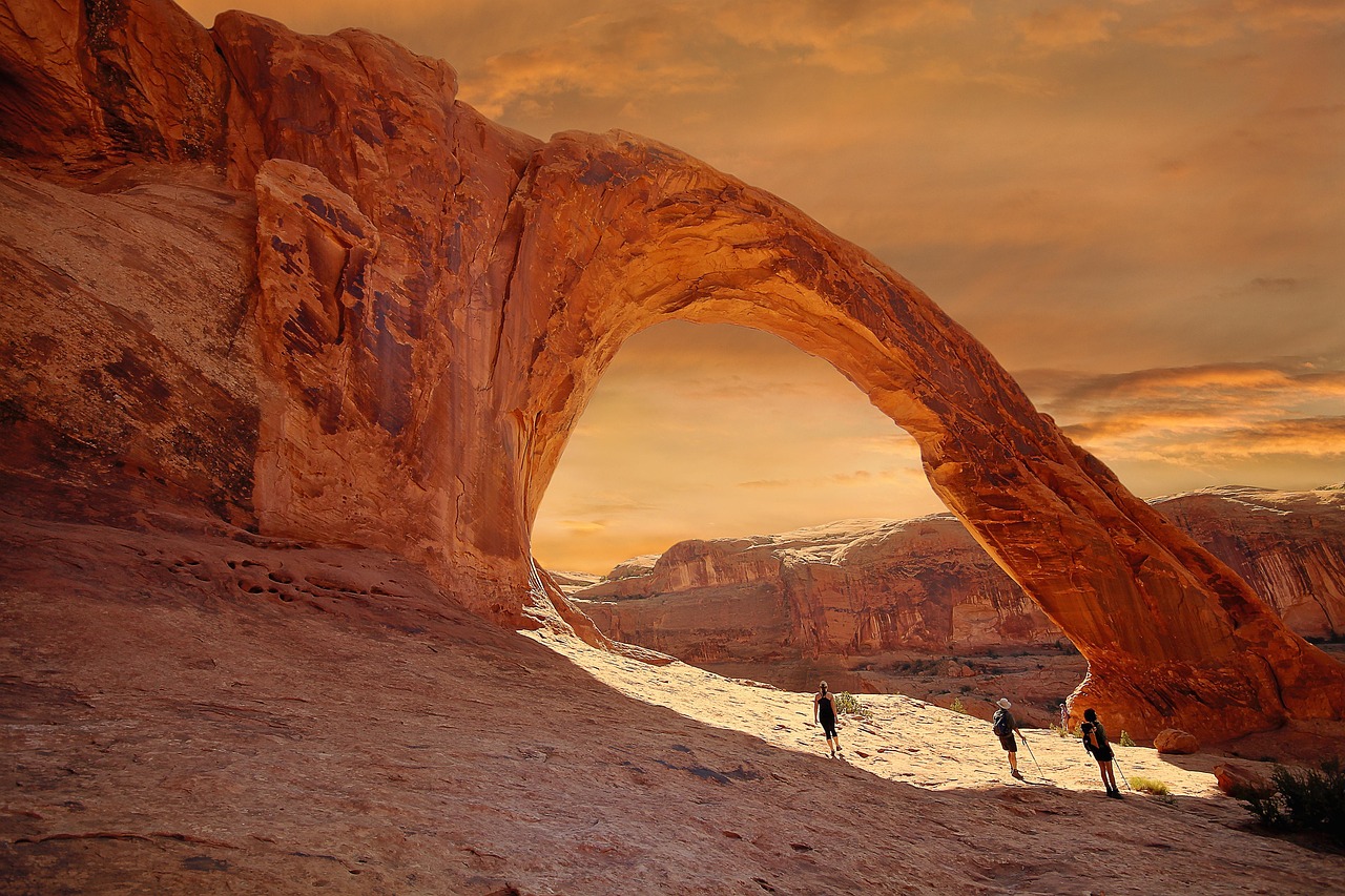 Corona Arch on BLM land outside Moab, Utah at sunset. Three hikers with hiking poles stand beneath, looking up at the arch.